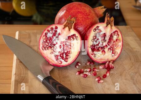 Nahaufnahme von einer ganzen und zwei halben Granatapfelfrüchten und einem damaszener Küchenmesser auf einem Holzschneidebrett mit Granatapfelkernen im Vordergrund Stockfoto