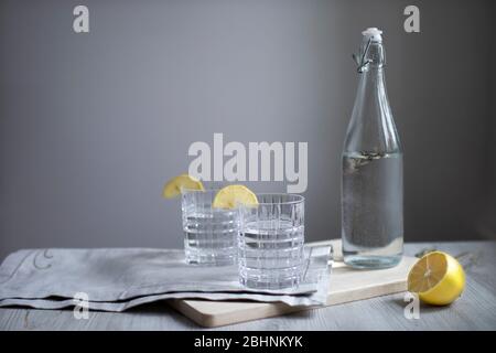 Zitronenlimonade mit Estragon in transparenter Flasche auf Tisch vor Hintergrund der grauen Wand. Zwei Wellgläser mit Zitronenscheiben. Graue Serviette und Stockfoto