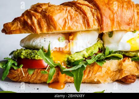 Croissant-Sandwich mit pochiertem Ei, Tomaten und Guacamole auf einem weißen Brett. Frühstück Essen Konzept. Stockfoto