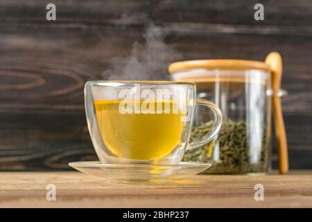 Glas transparente Tasse heißen grünen Tee mit Dampf und Dose mit Teeblättern und Löffel auf Holztisch gesundes Getränk, Antistress-Getränkekonzept Stockfoto