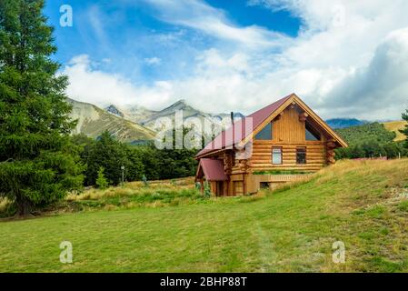 Berghütte. Log House. Solitary Cabin auf einem grünen Grasfeld mit Bäumen und einer Bergkette im Hintergrund. Stockfoto