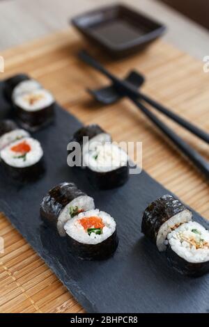 Maki Sushi auf einem schwarzen Brett. Sojasauce auf einem Teller und schwarze Essstäbchen auf einer Bambusmatte. Stockfoto