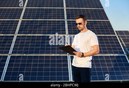 Porträt eines jungen Mannes, der an einem Solarpanel steht und in seinen Notizen nachdenkt, über einen Businessplan, das Konzept junger Geschäftsleute Stockfoto