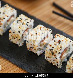 Uramaki serviert auf einem schwarzen Schiefer mit schwarzen Holzstäbchen und Sojasauce. Japanische traditionelle Küche mit Bambus-Service. Stockfoto