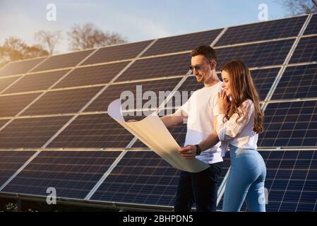Junge schöne Paar an einem sonnigen Tag steht bei einem Solarmodul und schaut in ein großes White Paper Plan, Gedanken über die Zukunft, Konzept der hellen Zukunft Stockfoto
