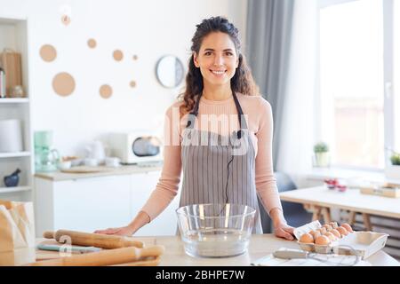 Horizontales mittleres Porträt einer fröhlichen jungen Frau, die Schürze trägt und am Küchentisch steht und die Kamera lächelt Stockfoto
