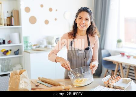 Fröhliche Food Blogger tragen Schürze stehen am Tisch in der modernen Küche schlagen Eier in Glas Schüssel mit Peitsche, Blick auf Kamera-Porträt Stockfoto