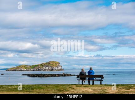 North Berwick, East Lothian, Schottland, Großbritannien. April 2020. UK Wetter: Ein ruhiger heller Tag. Trotz des ruhigen Wetters sind die Strände während der Sperrung fast leer. Ein Paar ruht sich auf einer Bank mit Blick auf den Firth of Forth und Craigleith Island in West Bay aus Stockfoto