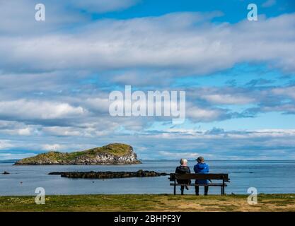 North Berwick, East Lothian, Schottland, Großbritannien. April 2020. UK Wetter: Ein ruhiger heller Tag. Trotz des ruhigen Wetters sind die Strände während der Sperrung fast leer. Ein Paar ruht sich auf einer Bank mit Blick auf den Firth of Forth und Craigleith Island in West Bay aus Stockfoto