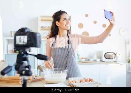 Attraktive junge Erwachsene Frau trägt Schürze, die Selfie nimmt, während sie Video für ihr Essen vlog Stockfoto