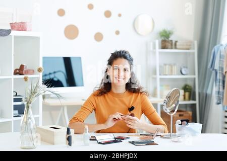 Fröhliche junge Erwachsene Frau am Tisch mit Beauty-Produkte auf sie sitzen Blick auf Kamera lächelnd Stockfoto