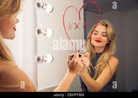 Eine Frau, die in einen Badezimmerspiegel schaut und mit Lippenstift ein Herz auf den Spiegel zieht. Stockfoto