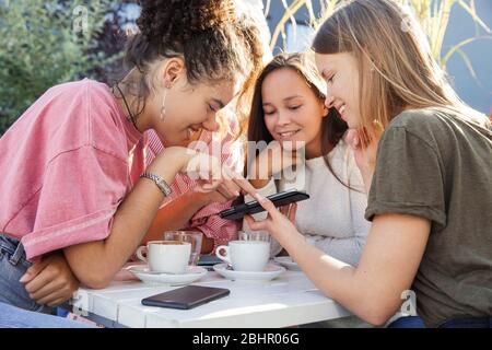 Drei Frauen sitzen um einen Tisch und schauen auf ein Handy und lächeln. Stockfoto