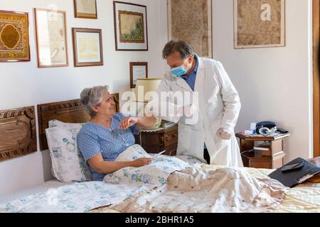 Ein Arzt in einem weißen Mantel und schützende Gesichtsmaske, die einen Hausbesuch bei einer älteren Frau Patientin, Begrüßung durch Berühren der Ellbogen. Stockfoto