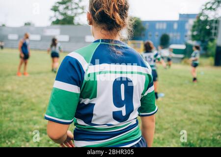 Rückansicht einer Frau, die ein blaues, grünes und weißes Rugby-Shirt auf einem Spielfeld beim Training mit anderen Spielern im Hintergrund trägt. Stockfoto