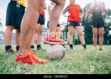 Die Beine und Füße einer Gruppe von Frauen, die auf einem Trainingsplatz stehen, eine mit einem Fuß auf einem Rugby-Ball. Stockfoto