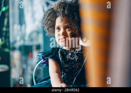 Ein Kind mit dunklem lockigen Haar in einem schwarzen Kleid mit einem Gesicht, das mit einer Spinne bemalt ist. Stockfoto