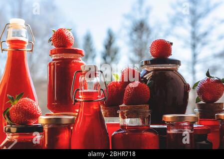 Hausgemachter Sirup und Marmelade, in Glasflaschen und Gläsern konserviert. Köstliche Erdbeersirup Flaschen in der Morgensonne. Viele Gläser mit Erdbeermarmelade Stockfoto