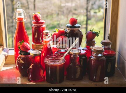 Viele Flaschen und Gläser mit hausgemachten Erdbeeren Sirup und Marmelade. Gläser Erdbeermarmelade auf der Fensterbank in der Morgensonne. Bio Stockfoto