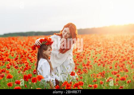 Kleine glückliche Mädchen mit Rotschopf Mutter in weißen Kleidern macht Kranz auf Mohn Feld bei warmen Sommer Sonnenuntergang Stockfoto