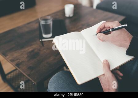 Aus einem hohen Winkel betrachtet, wie ein Mann auf dem Sofa sitzt und Notizen in Tagebuch oder Tagebuch auf seinen Beinen nimmt Stockfoto