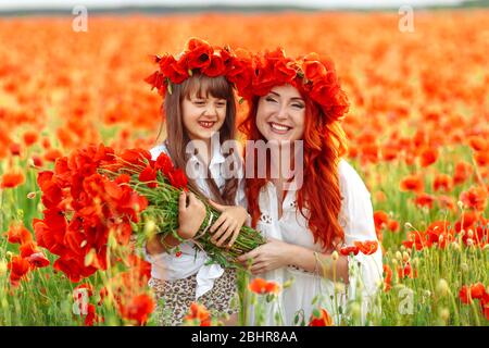 Kleines Mädchen mit Rotschopf Mutter in weißen Kleidern und Kranzen posiert mit Bouquet von Mohnblumen auf Mohnfeld bei warmen Sommer Sonnenuntergang Stockfoto