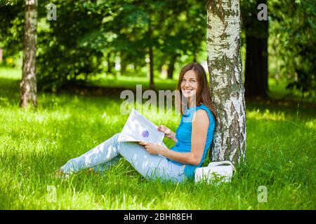 Junge schöne Brünette Frau liest ein Womens Magazin sitzt auf dem grünen Rasen im Sommerpark Stockfoto