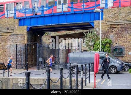 Zwei Männer joggen, ein Mann geht mit Anzug & Krawatte & Kopfhörer. DLR auf Brücke im Hintergrund in Limehouse Marina, Tower Hamlets, East London, Großbritannien. Stockfoto