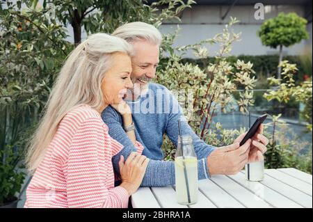 Ein Paar sitzt an einem Tisch im Garten mit Getränken in Flaschen, schaut zusammen auf ein Mobiltelefon. Stockfoto