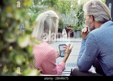 Rückansicht eines Paares, das in einem Garten sitzt und auf ein Mobiltelefon schaut. Stockfoto