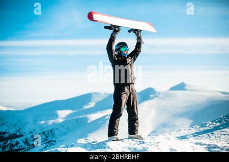 Eine Person, die einen schwarzen Skianzug, Helm und Brille auf einem Berg trägt und ein rotes Snowboard über dem Kopf hält. Stockfoto