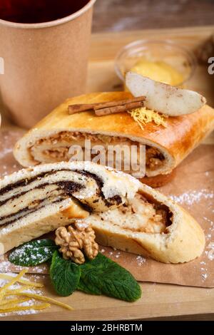 Tee in einer braunen Papiertasse, Mohn und Apfelstrudel auf einem rustikalen Holztablett. Nahaufnahme, verschwommener Hintergrund. Stockfoto