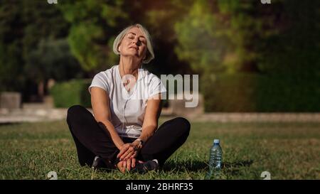 Reife Frau auf dem Gras sitzend, nach einem Lauf ruhend und Musik hörend Stockfoto