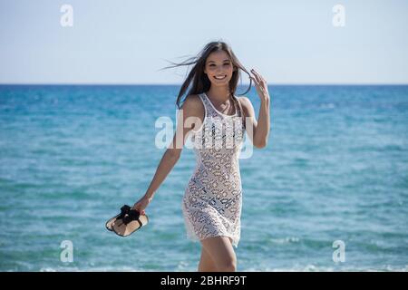 Junge Frau mit langen braunen Haaren, die am Meer spazieren und die Kamera lächeln. Stockfoto