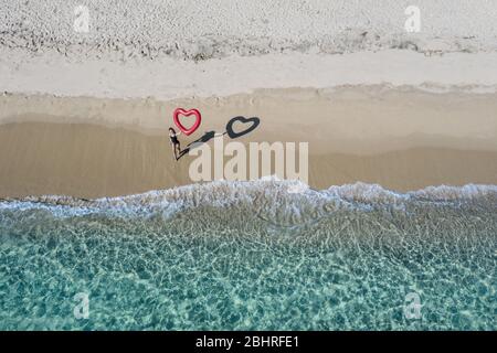 Luftaufnahme einer jungen Frau in Badeanzug, die an einem Sandstrand steht und einen roten, herzförmigen Ballon hält. Stockfoto