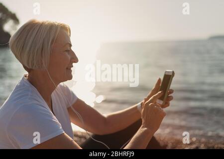 Reife Frau sitzt nach dem Joggen am Strand. Ältere Dame mit Smartphone und Kopfhörer bei Sonnenuntergang. Abends laufen Stockfoto