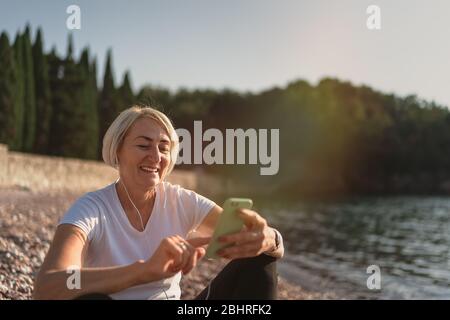 Reife Frau sitzt nach dem Joggen am Strand. Ältere Dame mit Smartphone und Kopfhörer bei Sonnenuntergang. Abends laufen Stockfoto