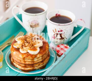 Stapel von Pfannkuchen mit Ahornsirup, gehackte Bananen und Pekannüsse. Zwei Tassen Kaffee auf dem Frühstückstablett Stockfoto