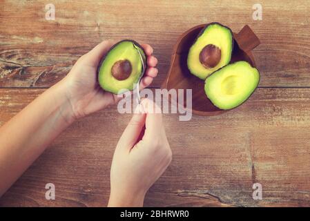 Weibliche Hände halten Löffel und die Hälfte der frischen reifen Avocado. Frau schält Avocado. Grüne Avocado auf Holztisch. Gesundes Öko-Essen. Produkt aus biologischem Anbau Stockfoto