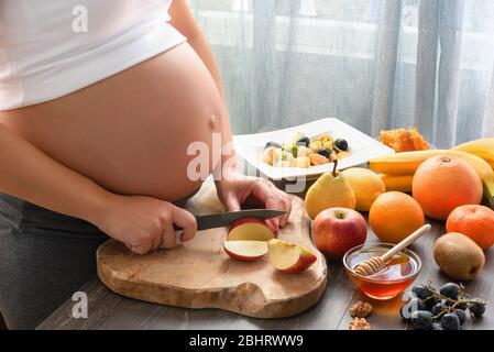 Junge und schöne schwangere Frau bereitet gesunde Obstsalat zu Hause. Bild von schwangeren Frau, die einen Obstsalat mit Kiwi und Banane kocht Stockfoto