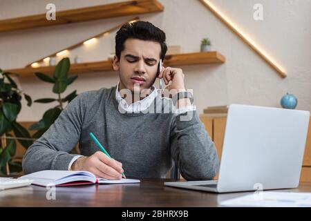 Geschäftsmann spricht auf Smartphone und Schreiben in Notebook in der Nähe Laptop am Tisch Stockfoto