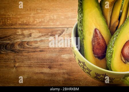 Nahaufnahme Foto von frisch geschnittenen Avocados, braune Samen sichtbar in grüner Schüssel auf braunem Holzhintergrund. Vegetarisches Speisekonzept, Vintage-Stil. Stockfoto