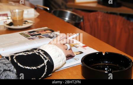 Die Hand des weiblichen Rauchers hält eine brennende Zigarre, während er eine Zeitschrift liest und Kaffee trinkt. Stockfoto