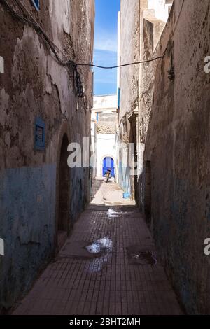 Schmale Straße voller Schatten, mit einem erleuchteten weißen Haus im Hintergrund. Hellblaues Tor mit einem Bogen und einem Motorrad vor dem Hotel. Essaouira, Moro Stockfoto