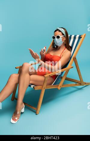 Frau in Sonnenbrille und medizinische Maske Handdesinfektionsmittel auf Liegestuhl auf blau auftragen Stockfoto