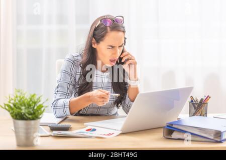 Wütende Frau, die in ein Mobiltelefon schreit und auf ein Notebook in einer Büroumgebung zeigt. Stockfoto