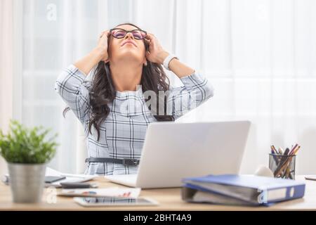 Büroassistentin, die sich auf ihrem Stuhl zurücklehnt und den Kopf in den Händen hält, da sie bei der Arbeit Kopfschmerzen hatte. Stockfoto