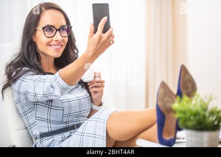 Gut aussehende Brünette Frau, die einen Kaffee hält, ein Selfie macht, ihre Füße in hohen Absätzen hat. Stockfoto