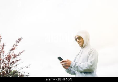 Junger Mann in grauem Sweatshirt und blauer Sonnenbrille, der das Handy in den Händen hält und mit Bäumen und weißem Hintergrund lächelt Stockfoto