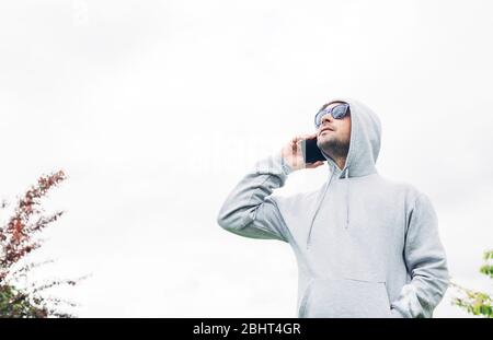 Junger Mann in grauem Sweatshirt und blauer Sonnenbrille, der das Handy in den Händen hält und auf weißem Hintergrund lächelt Stockfoto